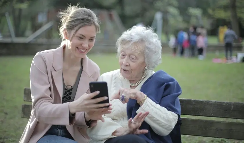 Smiling young woman showing something on a smartphone to an elderly woman on a park bench, with a playground and people in the background.