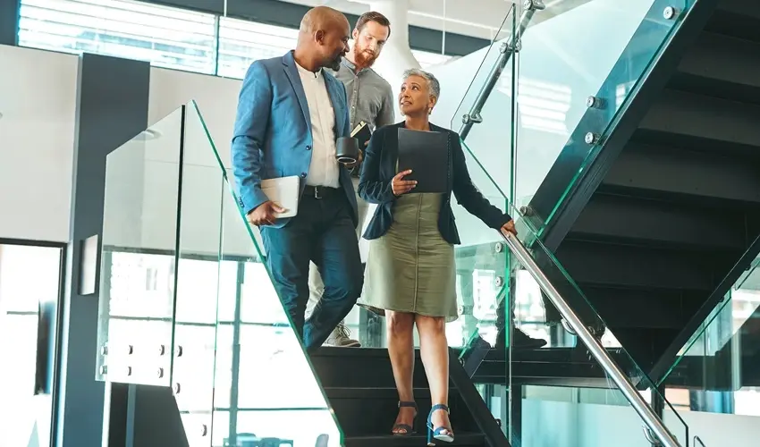 Three business professionals walking down glass stairs, engaged in conversation in a modern office setting.