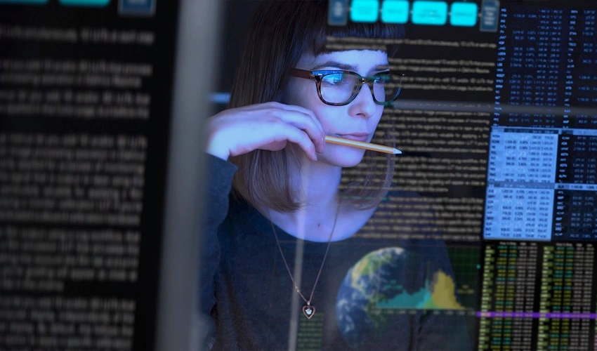woman with glasses biting pencil, looking at computer screen. the camera angle is behind the glass of the computer